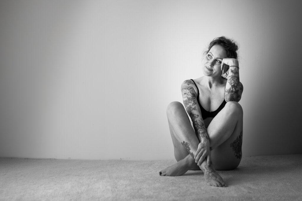 Schwarz-Weiß-Portrait einer tätowierten Frau, die im Studio auf dem Boden sitzt, den Kopf in die Hand gestützt und lächelnd zur Seite blickt